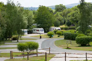 Wohnwagen auf einem Campingplatz, gepflasterter Weg und Hecken zwischen grünen Bäumen, im Hintergrund Hügel und Tal.
