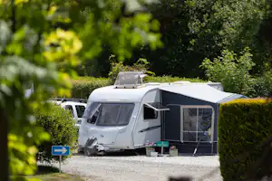 Wohnmobil und Wohnwagen auf einem Campingplatz, mit Vorzelt und Tisch am Stellplatz, umgeben von Hecken und Bäumen.