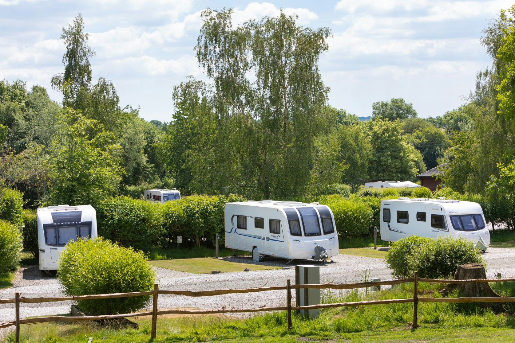 Wohnwagen auf einem Campingplatz-Stellplatz, umgeben von Hecken und Bäumen, mit Schotterweg und mehreren Fahrzeugen.