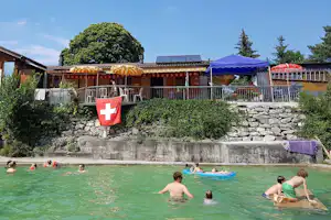 Außenpool auf dem Campingplatz mit Schwimmern, Sonnenschirmen und Liegen vor einem Gebäude und Hecken.