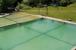 Schwimmbecken mit gelbem Geländer und Treppe am Rand, umgeben von Rasen, Bäumen und Liegen im Campingbereich.