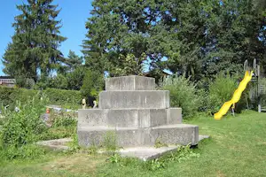 Betonstufen mit kleinem Wasserspiel, gelber Rutsche und Spielgeräten im Grünen zwischen Bäumen und Hecken.