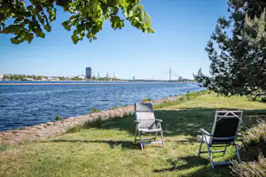 Zwei Campingstühle auf einer grünen Wiese am Seeufer, im Hintergrund Hafen und Brücke.