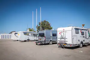Mehrere Wohnmobile auf dem Campingplatz-Parkplatz, mit Markise, Fahrradträger und Wäscheleine vor blauem Himmel.