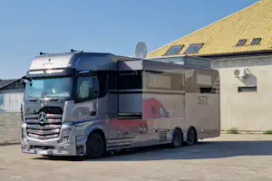 Silbernes Wohnmobil mit Markise und Satellitenschüssel an einer Hauswand, gelbes Ziegeldach und Fenster im Hintergrund.