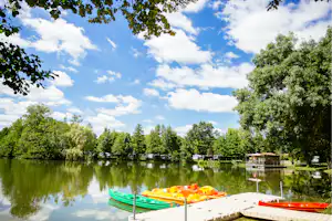 Ruderboote am Steg vor ruhigem See, im Hintergrund Bäume und kleine Gebäude am Campingplatz.