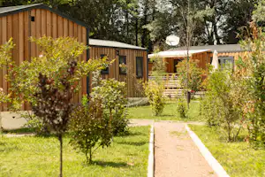Holzhäuser und Garten mit Rasen, schmalem Weg und Bäumen, im Hintergrund eine Terrasse mit Holzgeländer.