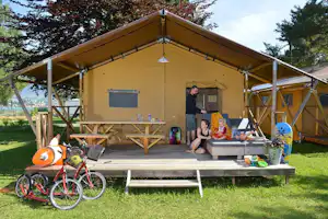 Familienzelt mit Holzveranda, Sitzbank und Spielzeug auf der Terrasse, umgeben von Rasen und Bäumen.