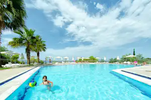 Schwimmbecken auf dem Campingplatz mit Palmen, Liegen und Sonnenschirmen am Rand, blaues Wasser und Himmel.