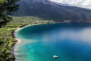 Bucht mit türkisfarbenem See, Boot im Wasser, Sandstrand und Blick auf bewaldete Berge und Ufergebäude.