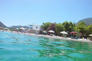 Blick auf das klare Wasser eines Außenpools am Campingplatz, mit Liegen, Sonnenschirmen und Gebäuden am Ufer.