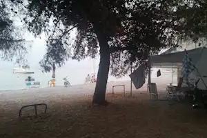 Campingplatz am See: Zelt mit Markise, Stühle und Tisch unter großen Bäumen, im Hintergrund Boote am Ufer.
