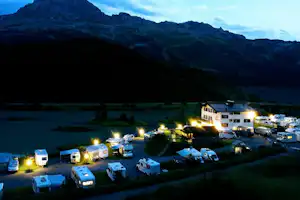 Campingplatz am See mit beleuchteten Stellplätzen, Wohnwagen und Zelten, im Hintergrund Berge und dunkle Uferlinie.