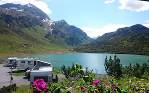 Wohnwagen und Campingfahrzeuge am Seeufer mit Blumenbeeten, im Hintergrund grüne Berge und ein breiter Stausee.