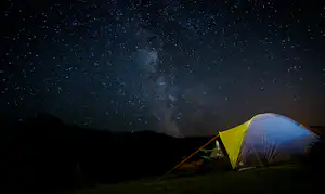 Campingzelt im Dunkeln unter der Milchstraße am Nachthimmel, mit Bergen im Hintergrund und Zeltbeleuchtung am Boden.