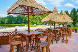 Holzterrasse mit Tischen und Stühlen unter Strohdächern, Blick auf den See und Boote am Strand im Campingbereich.
