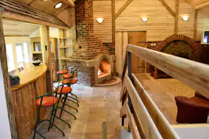 Innenbereich eines Holzhauses mit Kamin, Sitzbank, orangefarbenen Stühlen und Holztisch im warm beleuchteten Raum.