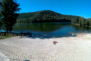 Sandstrand am See mit Paddlern auf dem Wasser, Bäume am Ufer und bewaldete Hügel im Hintergrund.