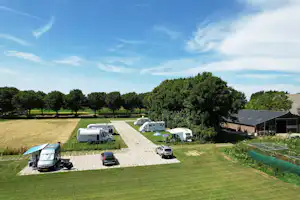Campingplatz mit Stellplätzen auf Rasen und gepflasterten Wegen, Wohnwagen und Autos, Bäume und ein Gebäude im Hintergrund.