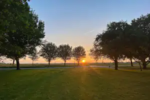 Grüne Wiese mit Bäumen am Campingplatz, im Hintergrund Felder und ein Sonnenuntergang am Horizont.
