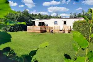 Wohnwagen mit Holzterrasse, Markise und Fahrrad vor Hecken und Rasenfläche im Campinggelände, umgeben von Bäumen.