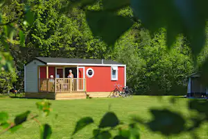 Rotes und weißes Holzhaus mit Veranda, davor ein Fahrrad auf dem Rasen, im Hintergrund Bäume.