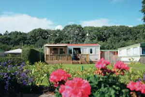 Mobilheim mit Holzterrasse und Markise, daneben ein weiterer Wohnwagen, umgeben von Rasen, Blumen und Bäumen.