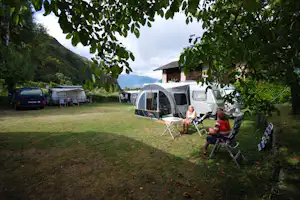 Campingplatz mit Vorzelt und Wohnwagen, Campingstühle am Tisch, Autos und weitere Zelte im Grünen unter Bäumen.