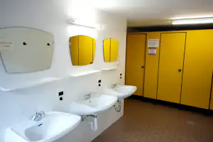 Camping-Badezimmer mit mehreren Waschbecken, weißen Fliesen und gelben Spinden, daneben Toilettenbereich mit Türen.