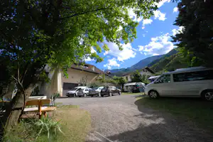 Parkplatz am Campingplatz mit mehreren Autos und Wohnmobilen, Kiesweg, Bäume und Berge im Hintergrund.