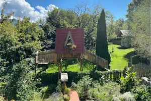 Spielhaus auf Stelzen mit rotem Dach im grünen Garten, umgeben von Hecken, Bäumen und Rasenfläche.