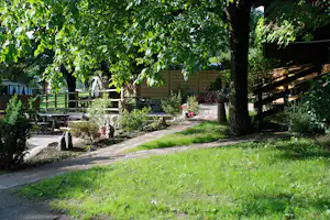 Holzhaus und Terrasse im Campinggarten mit gepflastertem Weg, Sitzbank und Blumenbeeten zwischen Bäumen.