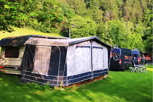 Wohnwagen mit Vorzelt und Markise auf dem Campingplatz, daneben ein Auto, im Hintergrund grüner Wald.