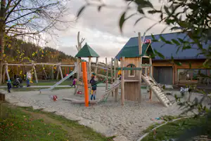 Spielplatz am Campingplatz mit Rutsche, Klettergerüst und Sandfläche, umgeben von Bäumen und Holzzäunen.