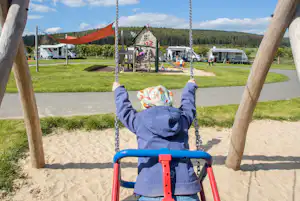 Blick auf eine blaue Kinderschaukel auf einem Sandspielplatz, im Hintergrund Wohnwagen und Wohnmobile auf dem Campingplatz.
