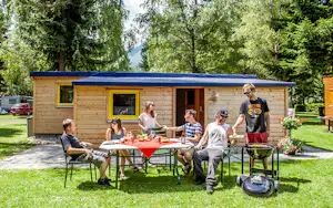 Familie am Esstisch vor einem Holz-Campinghaus mit Markise, im Garten mit Rasen und Bäumen.