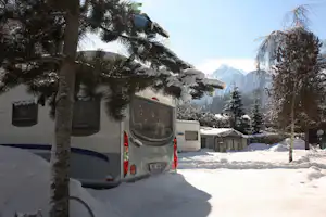 Wohnmobil im Schnee zwischen verschneiten Bäumen, im Hintergrund Berge und Zelte auf dem Campingplatz.