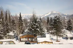 Verschneiter Campingplatz mit Wohnwagen und Zelten, Tannen und Bergen im Hintergrund.