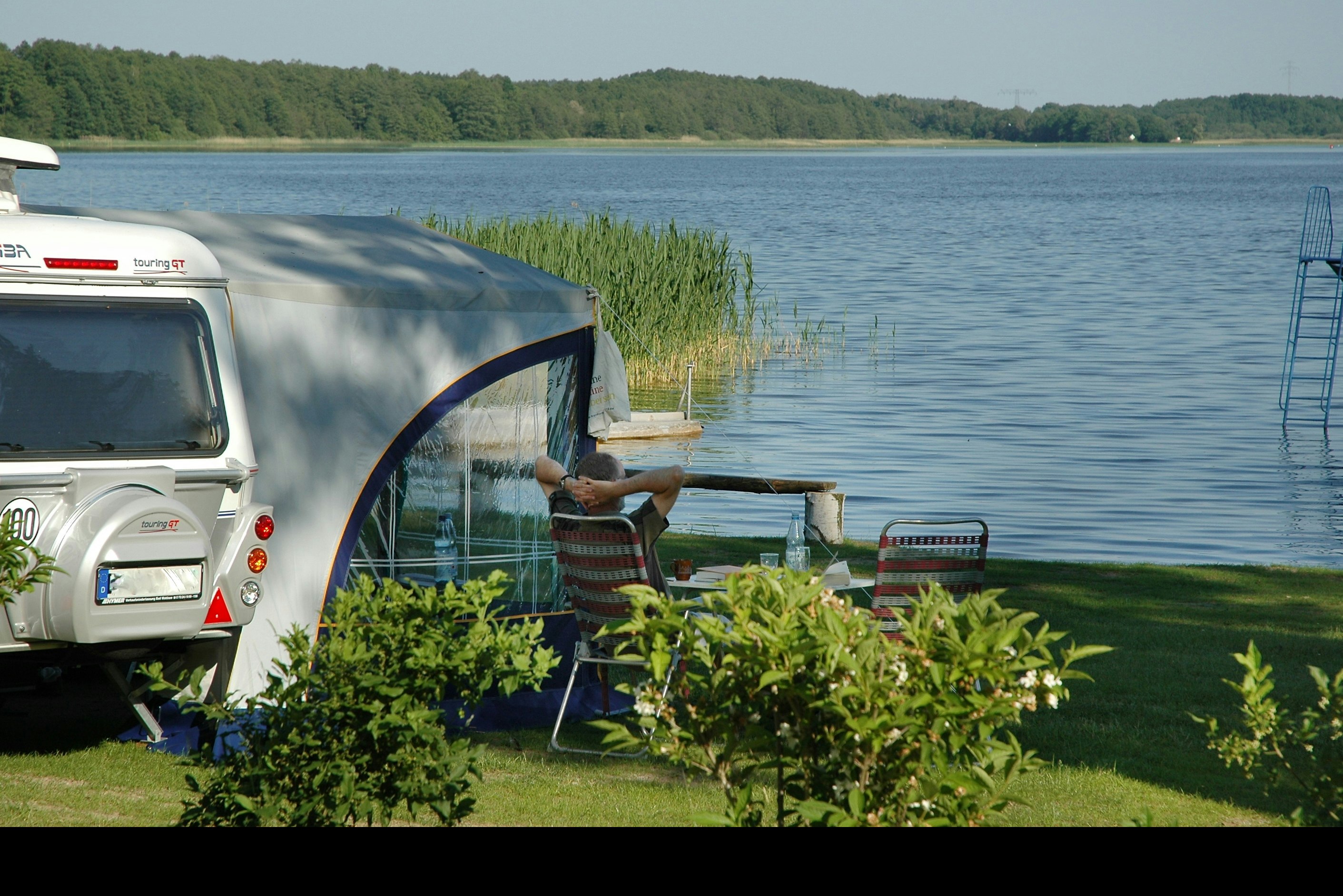 Wohnmobil am Seeufer mit Sitzgelegenheit und Pflanzenbeeten, im Hintergrund Wasser und bewaldete Hügel.
