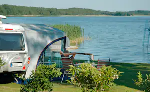 Wohnmobil am Seeufer mit Sitzgelegenheit und Pflanzenbeeten, im Hintergrund Wasser und bewaldete Hügel.