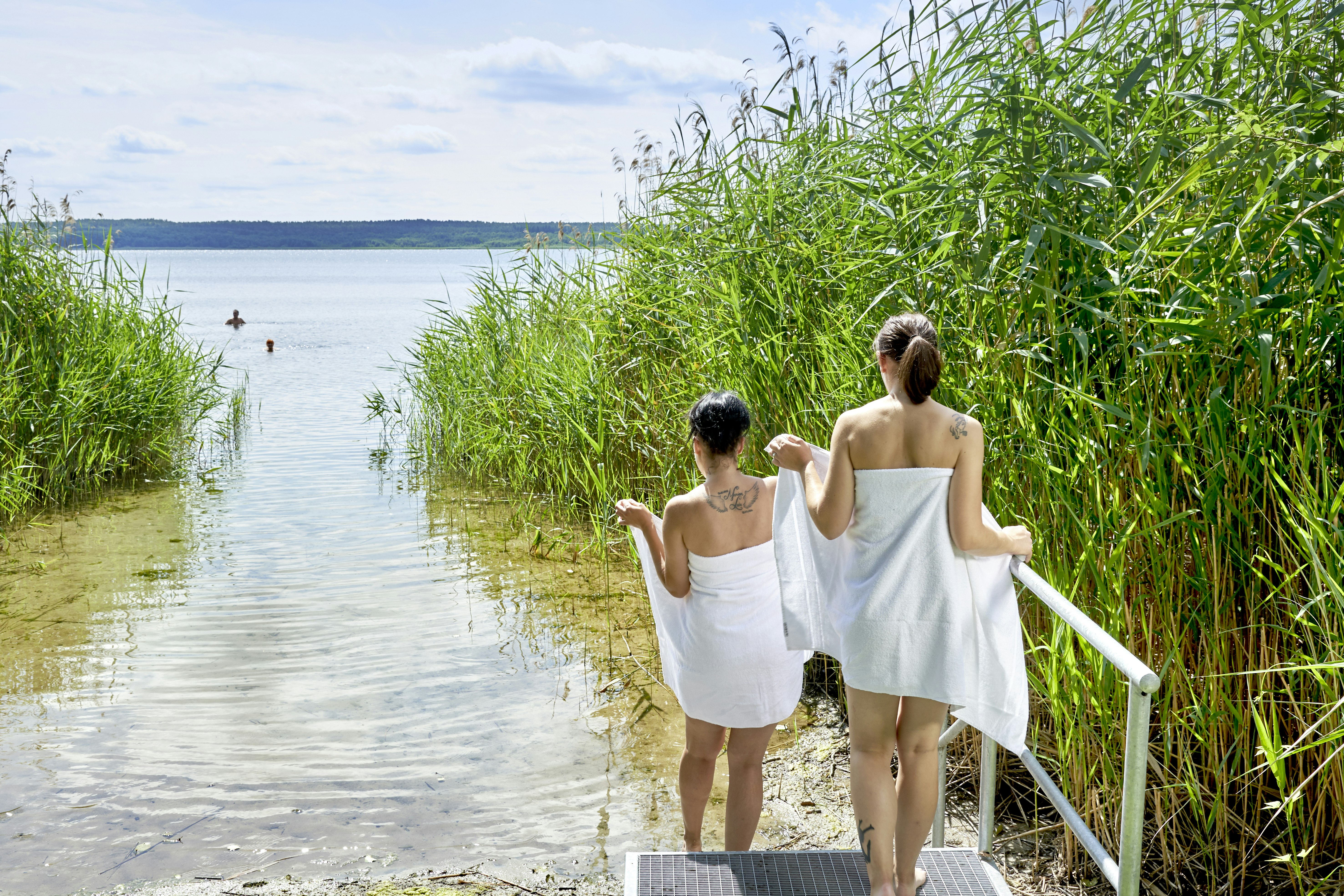 Zwei Personen am Seeufer im Schilf, mit Handtüchern und Bademänteln, Blick auf das Wasser und die entfernte Uferlinie.