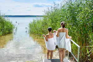 Zwei Personen am Seeufer im Schilf, mit Handtüchern und Bademänteln, Blick auf das Wasser und die entfernte Uferlinie.