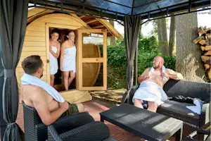 Holzsauna auf einer Camping-Terrasse, davor Liegen und Handtücher, im Hintergrund zwei Personen und ein Mann im Ruheraum.