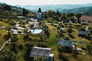 Campinganlage mit rundem Pool, vielen Stellplätzen, Zelten und Wohnmobilen inmitten von grünen Wiesen und Bäumen.