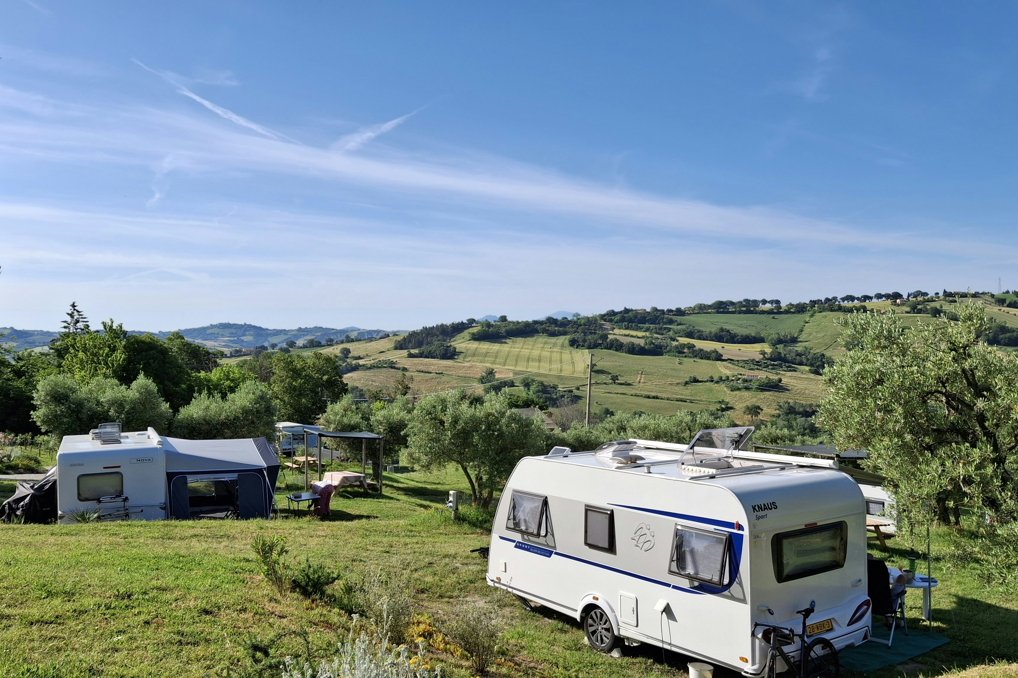 Wohnwagen und Zelt auf grünem Campingplatz am Hang, mit Bäumen, Gartenmöbeln und Blick über die Landschaft.
