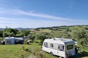 Wohnwagen und Zelt auf grünem Campingplatz am Hang, mit Bäumen, Gartenmöbeln und Blick über die Landschaft.