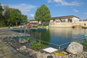 Überdachter Camping-Pool mit Metallgeländer, Steg und Steinbeckenrand, umgeben von Kieswegen und Bäumen.