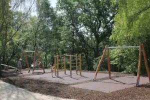 Spielplatz am Campingplatz mit Schaukeln und Klettergerüst auf gepflastertem Boden, umgeben von Bäumen im Grünen.