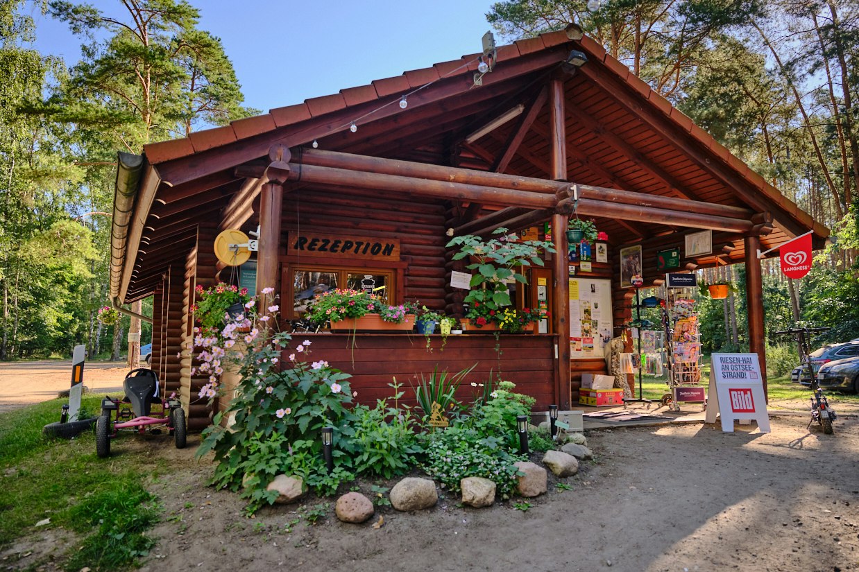 Holzgebäude mit Schild „Rezeption“ und Eingang, davor Blumenbeete und Weg, im Hintergrund Bäume und ein geparkter Wagen.
