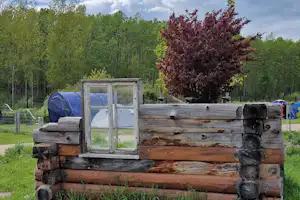 Holzunterkunft mit Fensterfront auf einem Campingplatz, im Hintergrund Zelte, Bäume und ein Zaun.